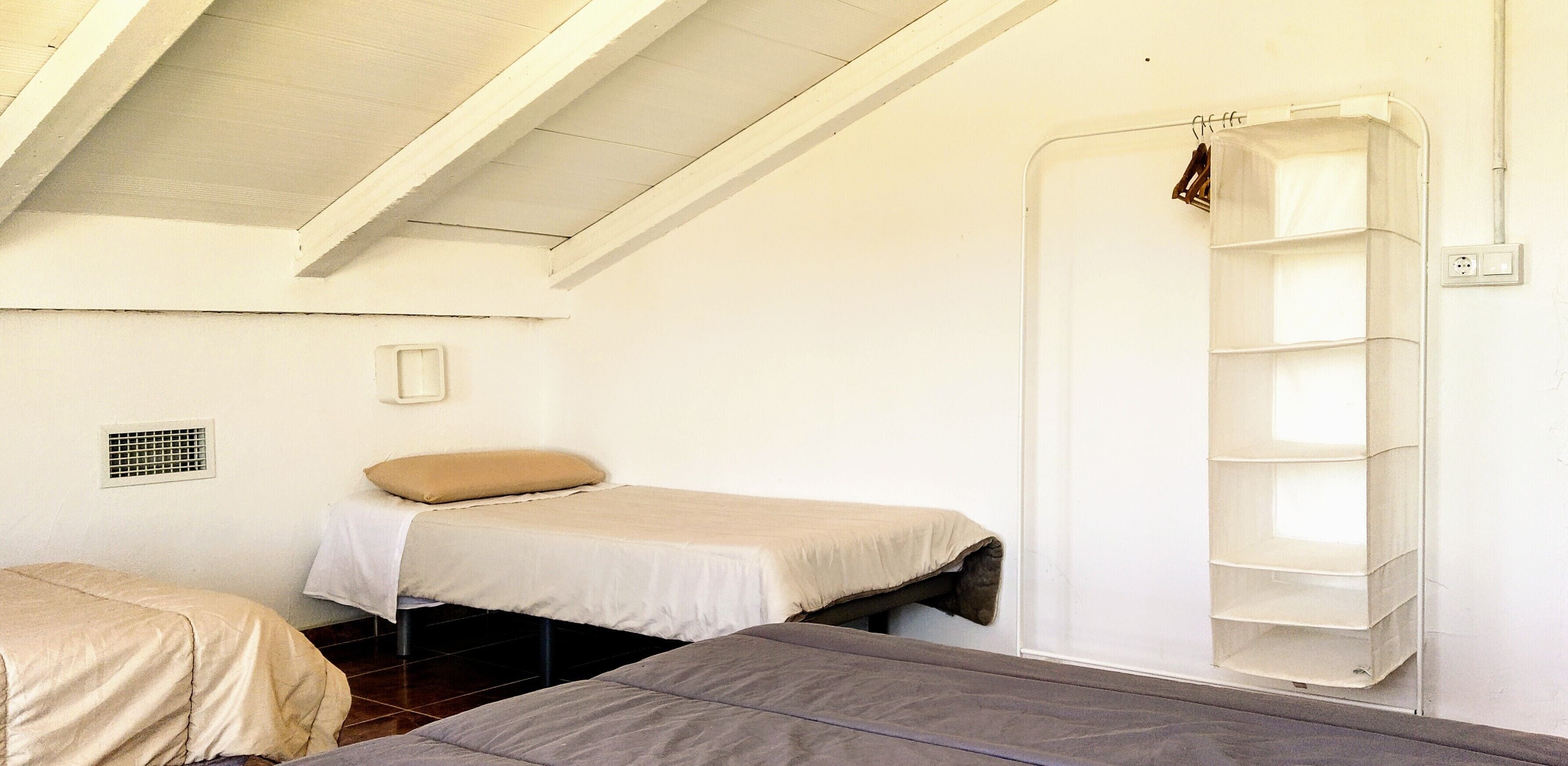 A small, simple room with two beds, a white shelf unit, and a vent on the wall.