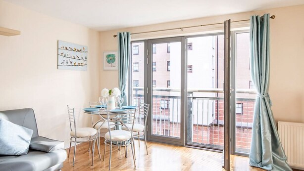 A modern living room with a grey sofa, a dining table with chairs, a balcony with a view of buildings, and a sliding glass door.