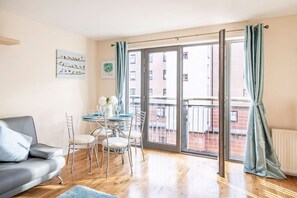 Un salon moderne avec un canapĂ© gris, une table Ă manger avec des chaises, un balcon offrant une vue sur les immeubles et une porte-fenĂȘtre coulissante.