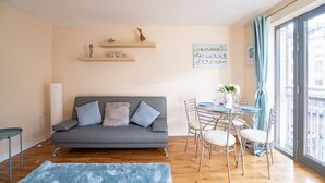 A living room with a grey sofa, a dining table with chairs, a blue rug, and a window with a view.