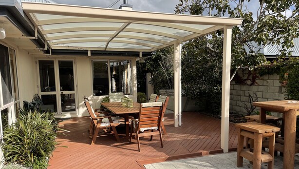 Un patio con una mesa y sillas de madera, una zona cubierta con techo de vidrio y un camino de ladrillo.
