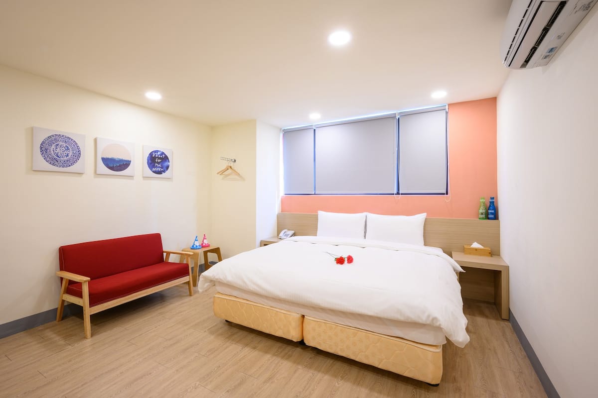 A hotel room with a bed, a red sofa, a bedside table, and a wall-mounted air conditioner.
