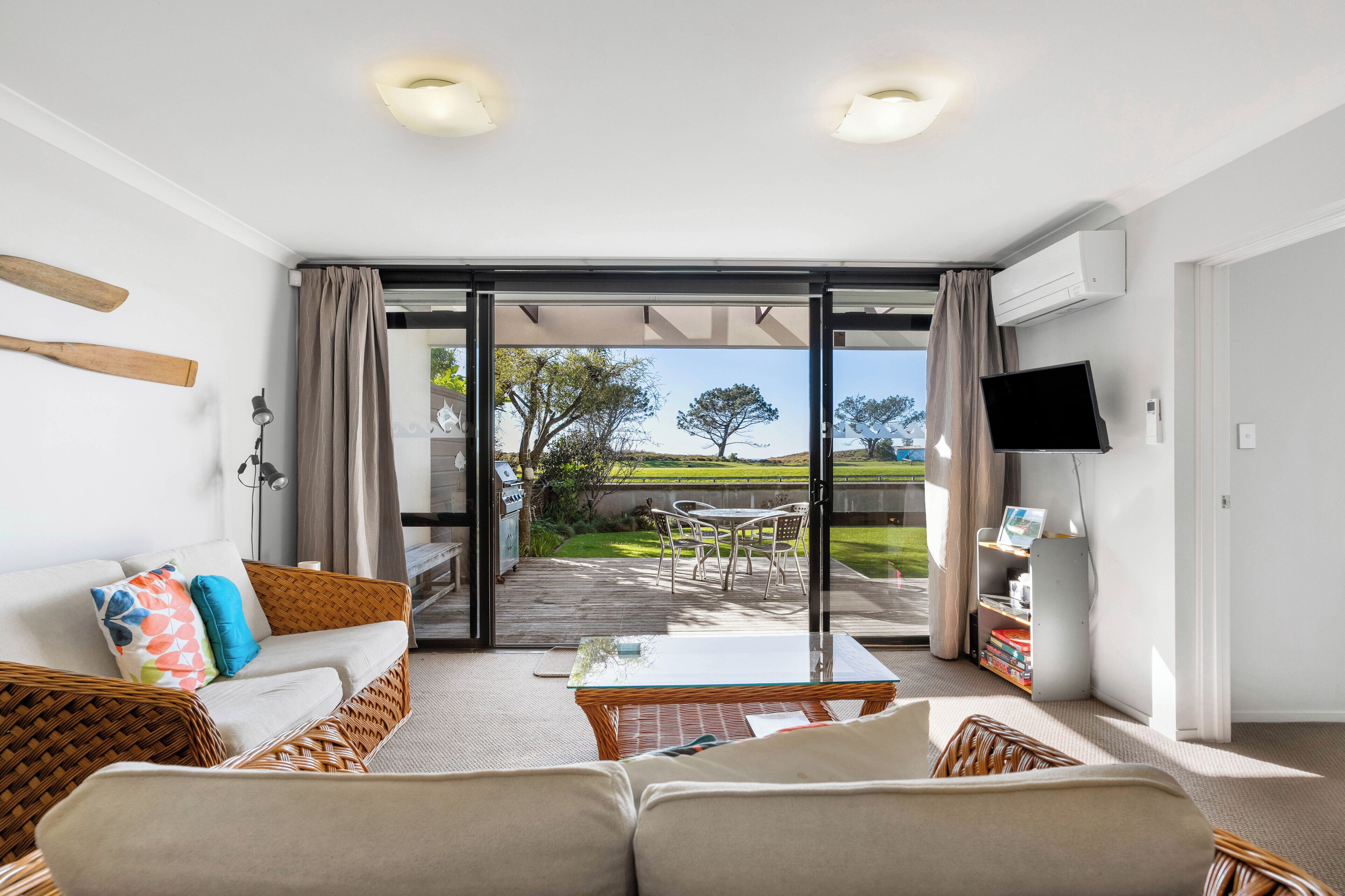 Ein modernes Wohnzimmer mit Sofa, Couchtisch und Fernseher. Eine Glastür führt auf eine Terrasse mit Grill und Blick auf eine grüne Landschaft.