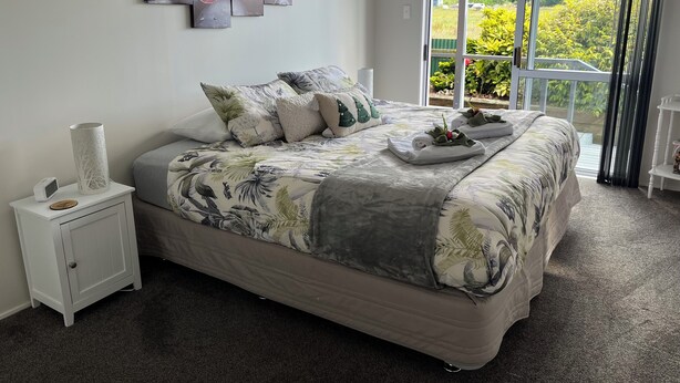 A bedroom with a large bed, a nightstand, a lamp, a painting, and a view of greenery through a sliding door.