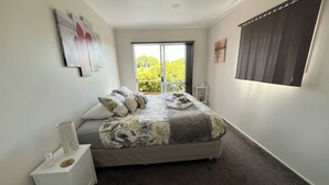 A bedroom with a bed, a nightstand, a window with blinds, and a view of greenery outside.