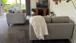 A living room with a grey sofa, a white throw blanket, a flat-screen TV, and a large window with a view of greenery.