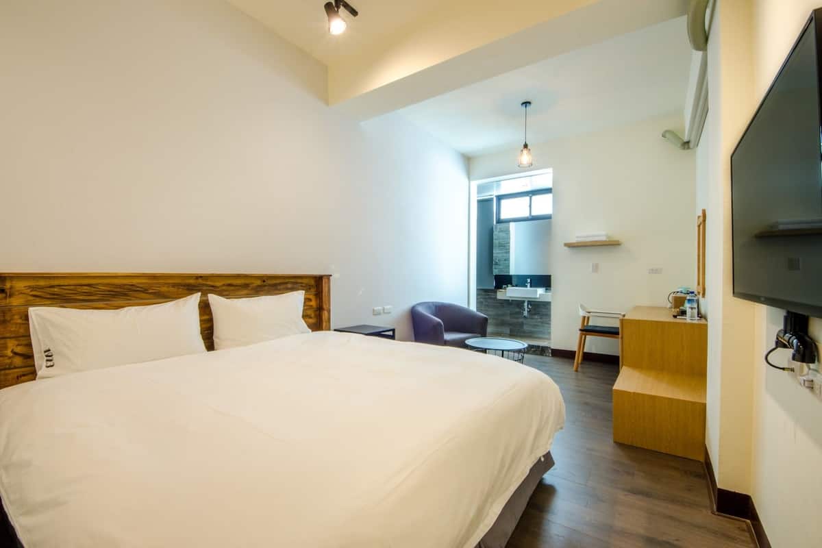 A hotel room with a large bed, a desk, a chair, a small table, a television, and a bathroom visible through an open door.
