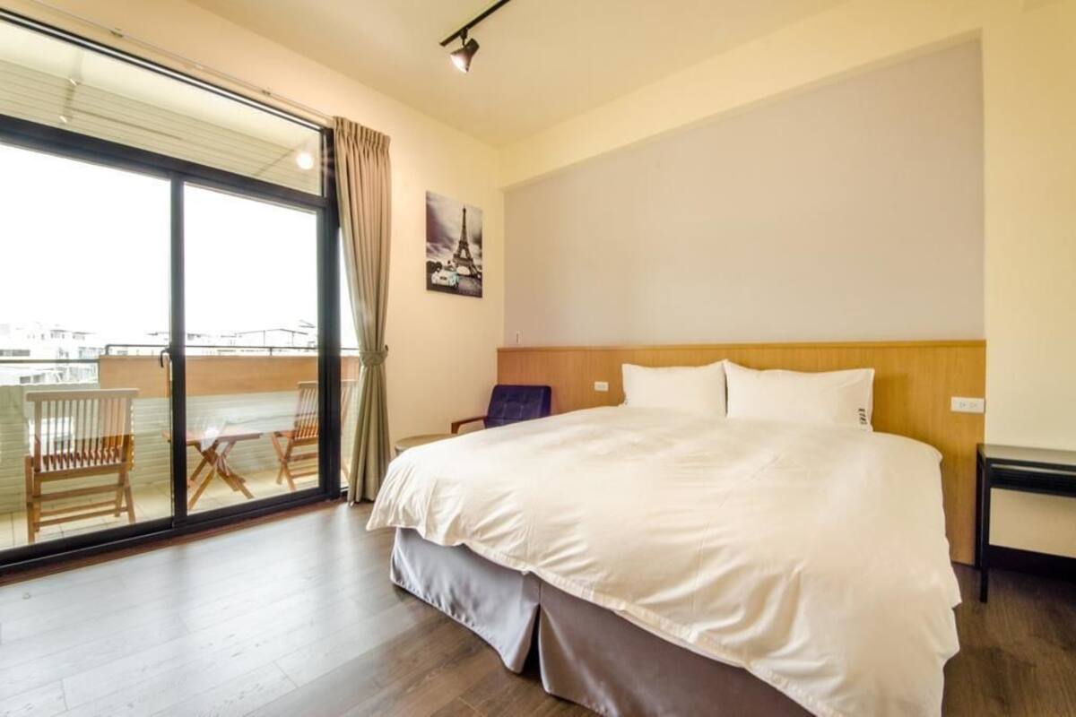A hotel room with a large bed, a wooden headboard, a view through a sliding glass door to an outdoor seating area, and a single chair.