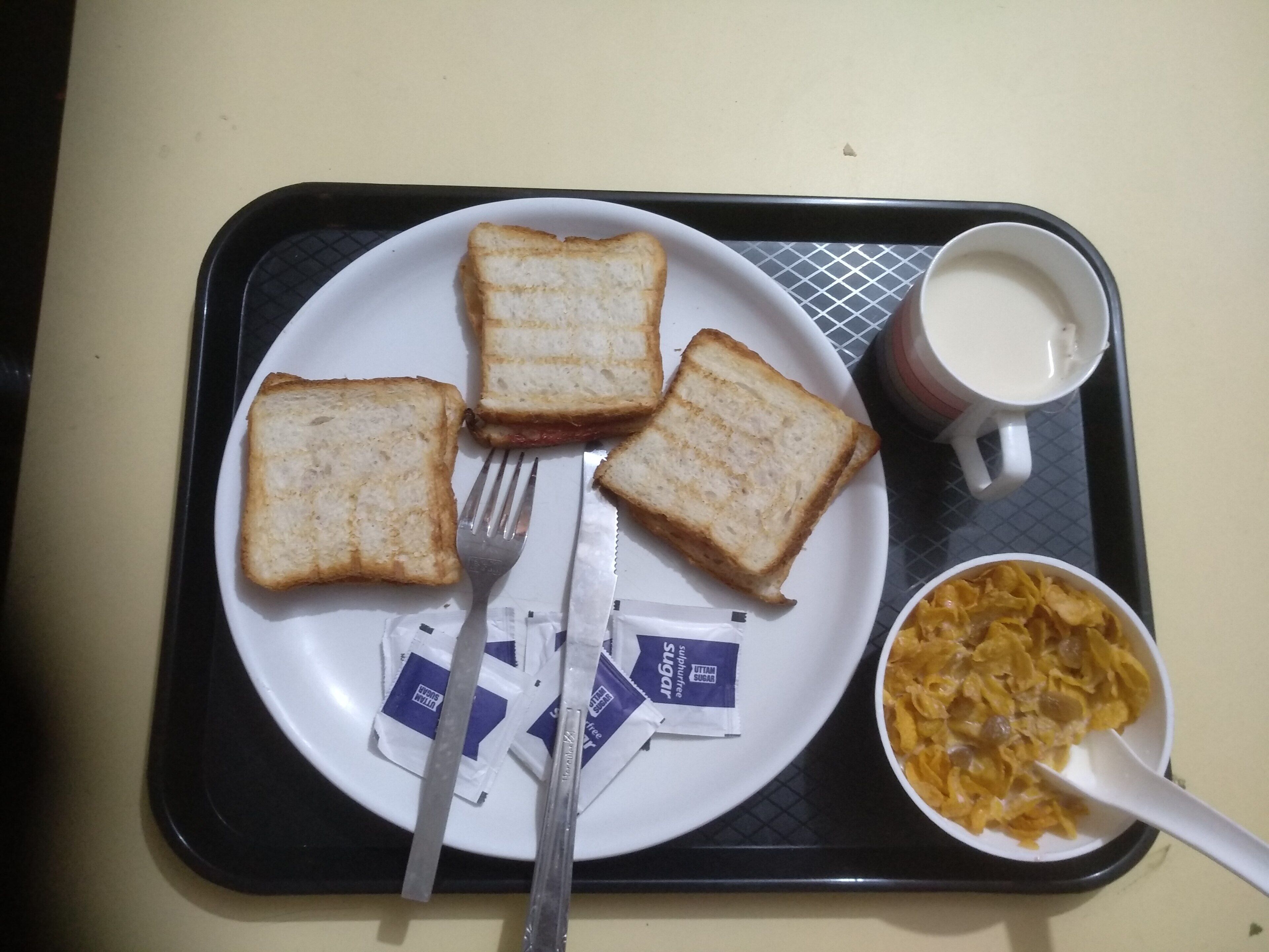 Ein Tablett mit einer Tellerviertel weiße Brotscheiben, einer Gabel, einem Messer, einer Tasse Milch und einer Schüssel mit Müsli und einem Löffel.