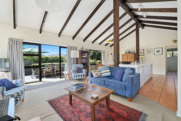 A spacious living area with a blue sofa, a wooden coffee table, and a red patterned rug. There is a large window with a view of greenery and mountains.