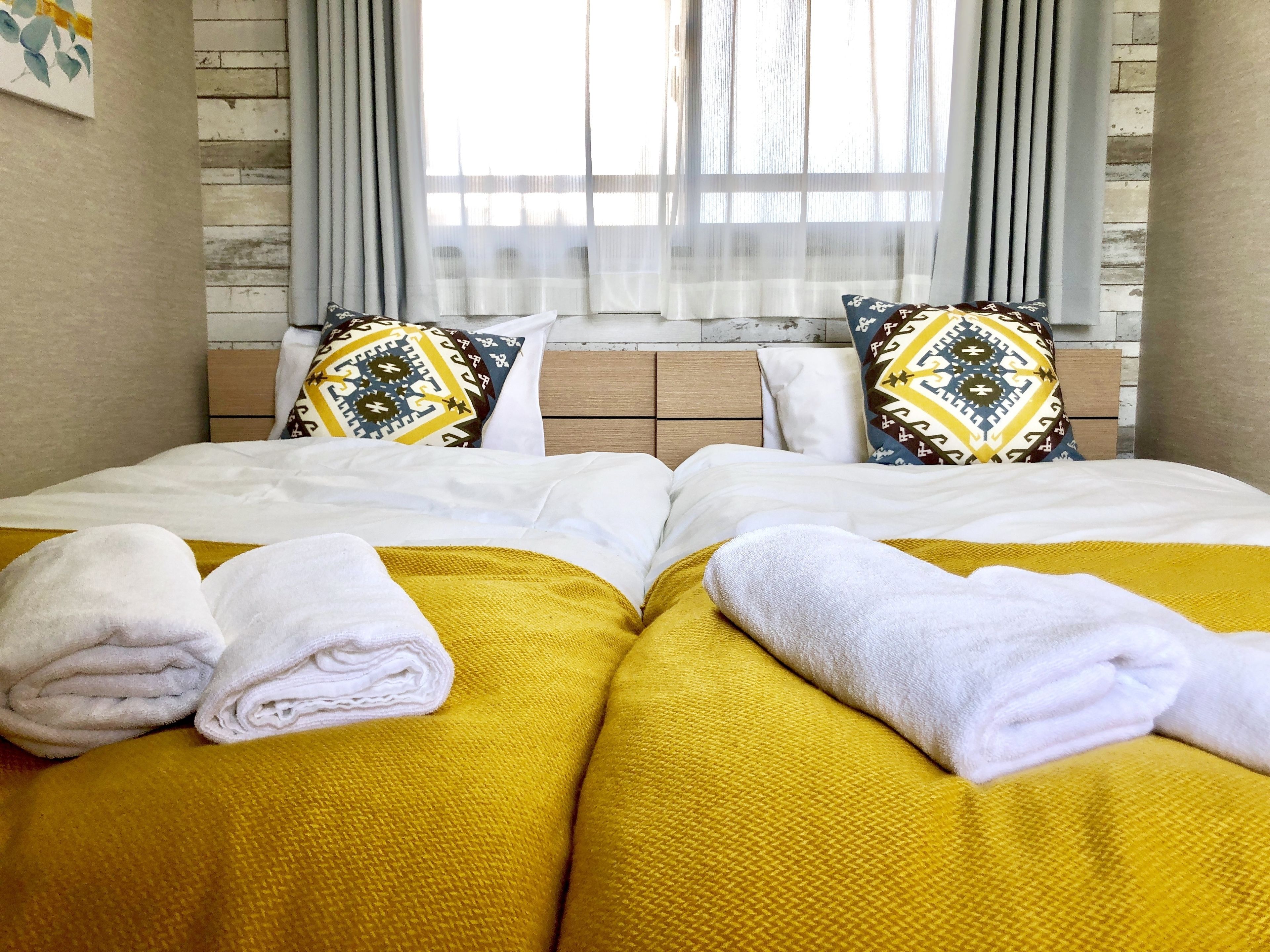 A hotel room with two beds, each with a yellow bedspread and white linens. There are decorative pillows with geometric patterns, and the room has a wooden headboard and a window with sheer curtains.