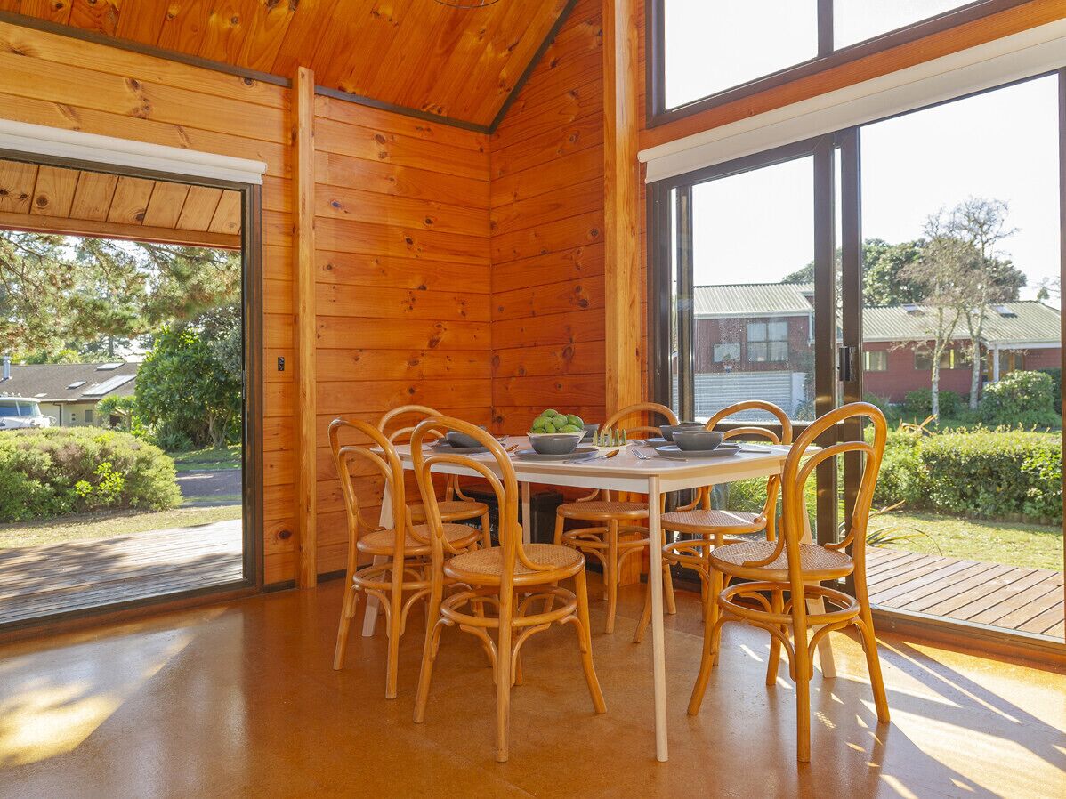 Een houten interieur met een eettafel en stoelen, grote ramen en uitzicht op buiten.