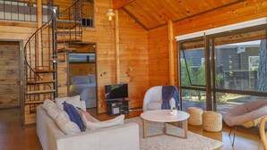 Interior acolhedor de uma cabana com decoração em madeira, escada, sofá, televisão e uma grande janela com vista para o exterior.