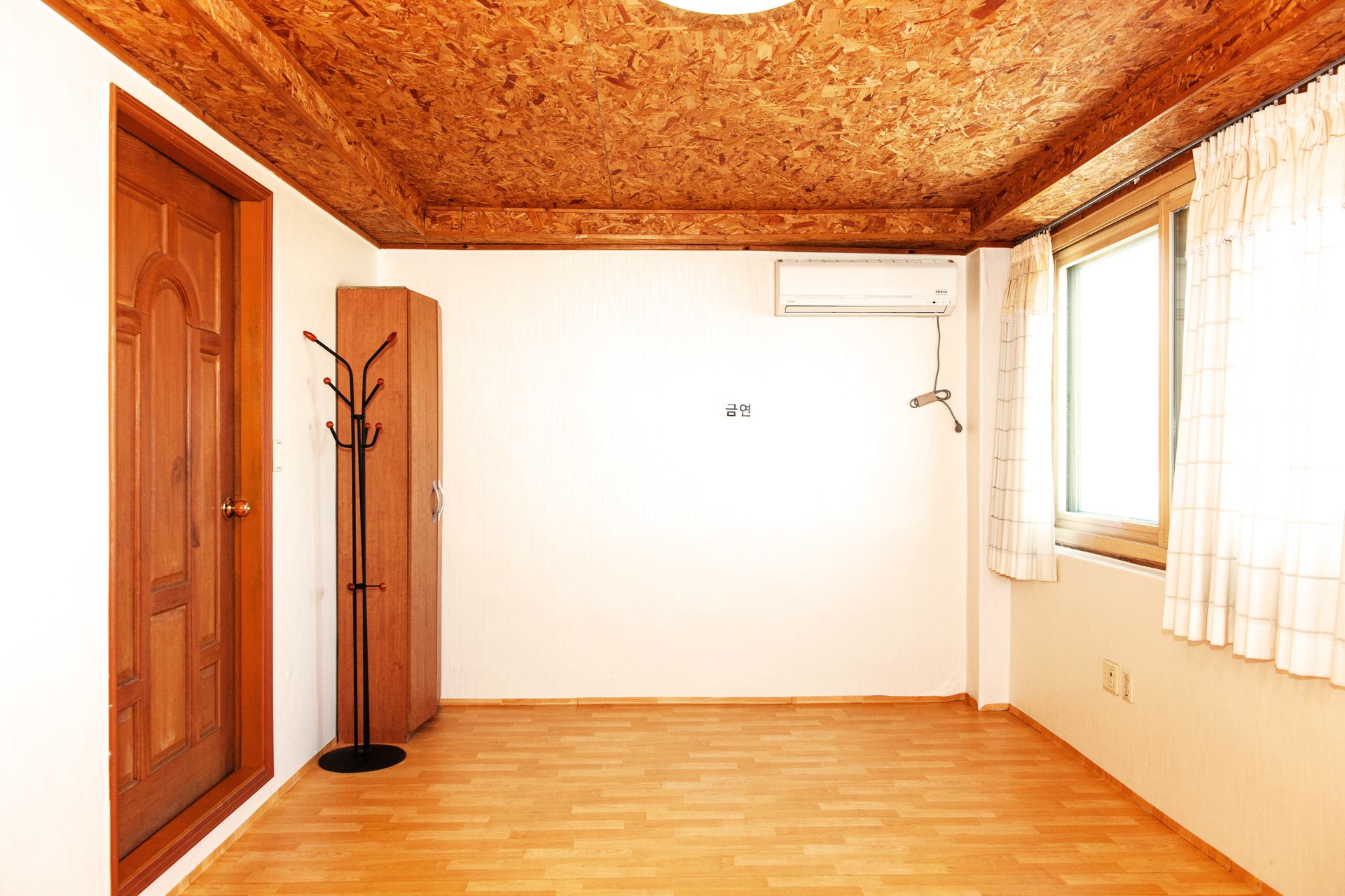 A room with a wooden floor, a wooden door, a coat rack, a window with curtains, and a wall-mounted air conditioning unit.