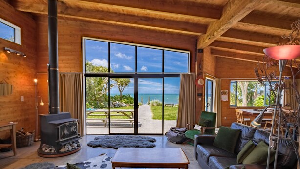 L’intérieur chaleureux d’un chalet avec un grand plafond en bois, une cheminée en pierre, des meubles en cuir et une vue sur la plage grâce à de grandes fenêtres.