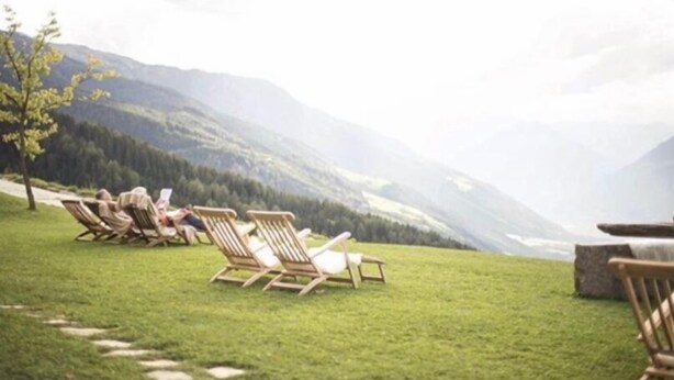 A row of wooden lounge chairs on a grassy lawn with a mountainous backdrop. - Tholer (Lagundo)