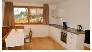 A modern kitchen with wooden flooring, a dining table with a white tablecloth, a window with curtains, and a view of a mountainous landscape. - Tholer (Lagundo)