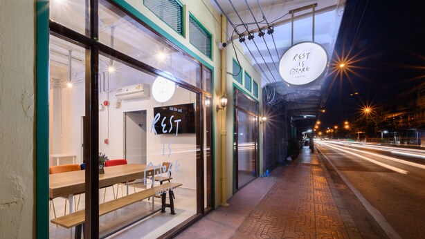 A hostel with a wooden bench outside, a sign reading "REST IS MORE HOSTEL", and a patterned sidewalk. - REST IS MORE Hostel (Bangkok)