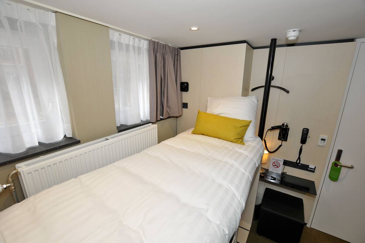 A hotel room with a bed, a radiator, a window with curtains, a bedside table with a phone and a sign, and a door with a green handle.