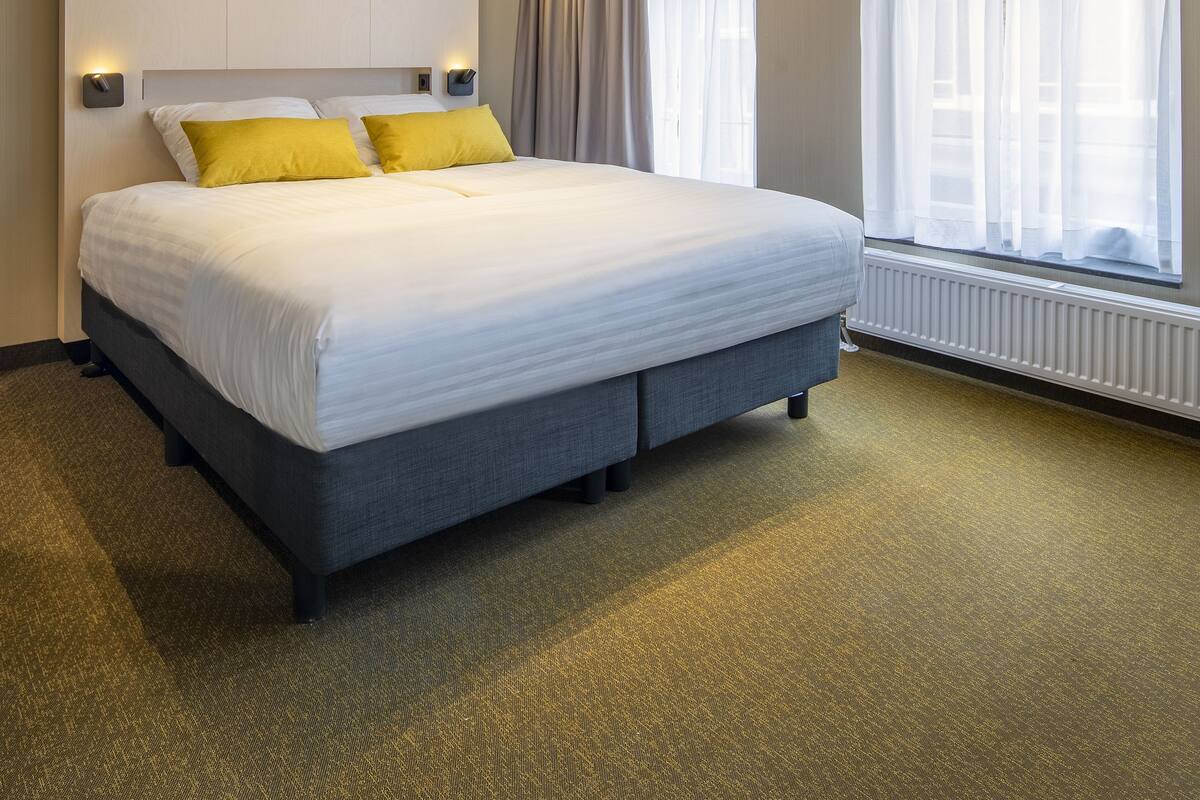A modern hotel room with a large bed, built-in headboard, and a view of the city through large windows.