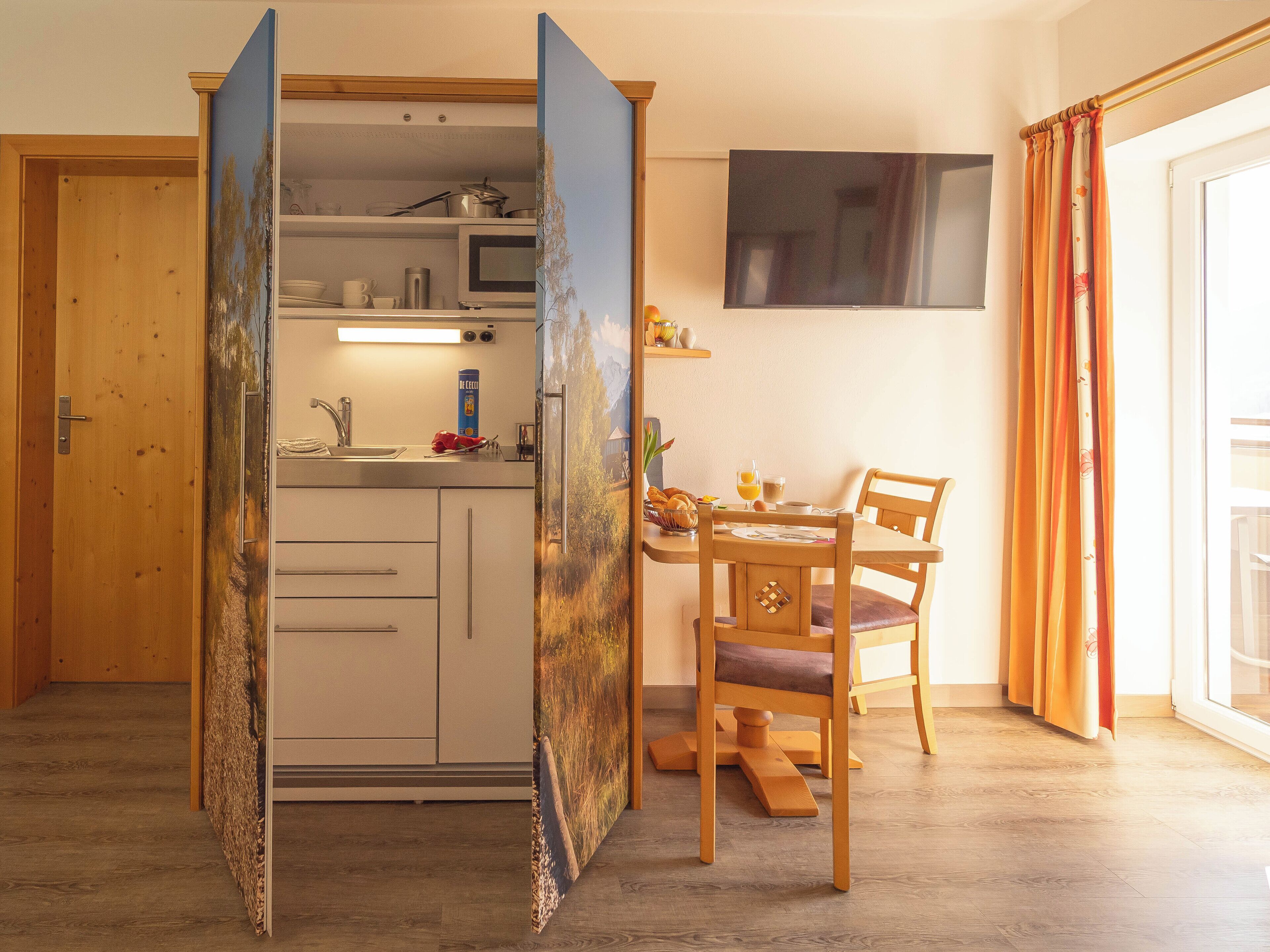 A compact kitchen with a microwave, sink, and cabinets. There's a dining area with a table and chairs, and a TV mounted on the wall.