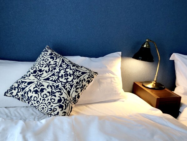 A neatly made bed with white linens and a single decorative pillow with a blue and white pattern. A black desk lamp is positioned on a wooden nightstand beside the bed.