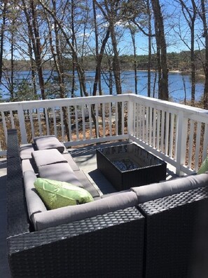 A deck with a sofa and a coffee table, surrounded by a white railing, overlooking a body of water and trees.