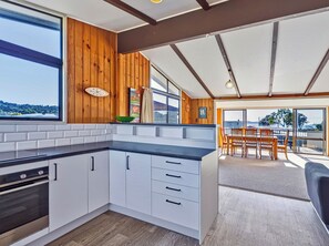 A modern kitchen with white cabinets, a black countertop, and wooden accents. There is a dining area with a table and chairs, and a view of the outdoors. - Joes Place - Cooks Beach Holiday Home (Cooks Beach)