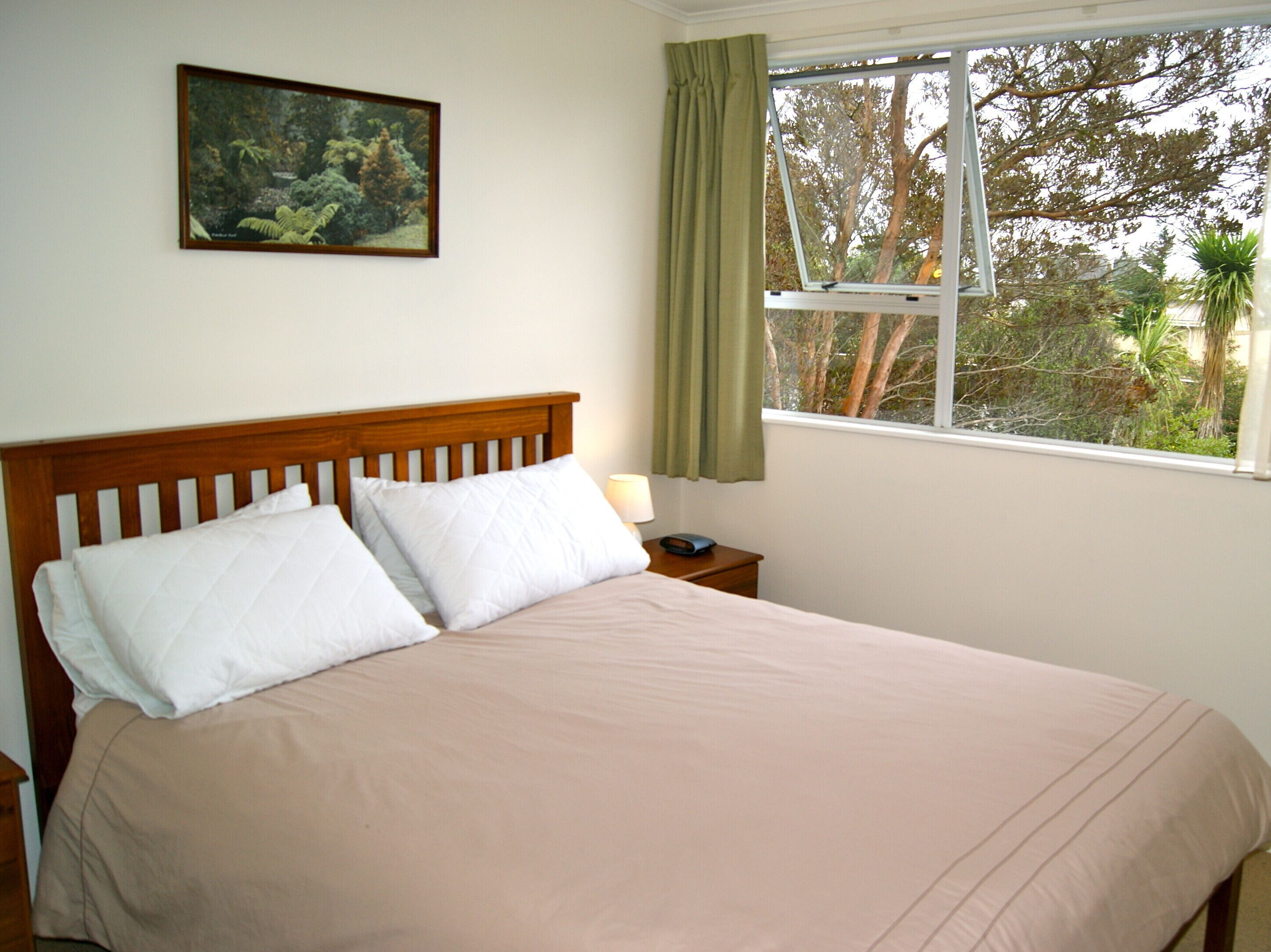 Une chambre à coucher avec un lit, une table de chevet, une lampe, une fenêtre avec des rideaux et un tableau encadré au mur.