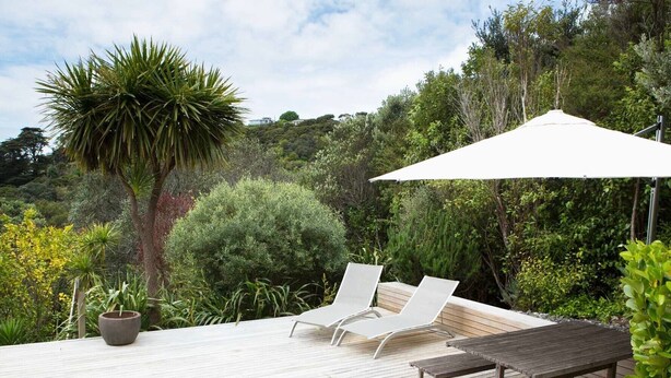 Een houten terras met twee lounge fauteuils en een tafel, omgeven door weelderige begroeiing en een grote parasol.