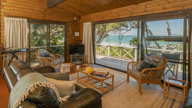 A wooden interior with a leather sofa, wicker chairs, a television, and a balcony with a view of the beach. - Norma's Place - Simpsons Beach Bach (Whitianga)