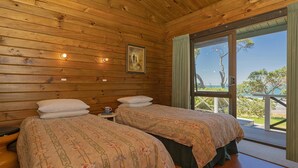 A cabin with two beds, wooden walls, a painting, and a balcony with a view. - Norma's Place - Simpsons Beach Bach (Whitianga)