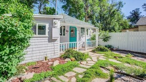 Ein kleines, eingeschossiges Haus mit einer Veranda, umgeben von einem Garten mit trittsteinförmigen Wegen und verschiedenen Pflanzen.