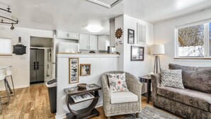 Ein modernes Wohnzimmer mit einem grauen Sofa, einem Korbstuhl und einem Holztisch. Im Hintergrund ist eine Küche mit weißen Schränken und einem eingebauten Backofen zu sehen.