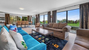 Ein geräumiges Wohnzimmer mit einem blauen Sofa, einem braunen Sofa und einem Couchtisch. Ein Essbereich mit Tisch und Stühlen lädt zum Verweilen ein. Große Fenster bieten einen Blick ins Freie.