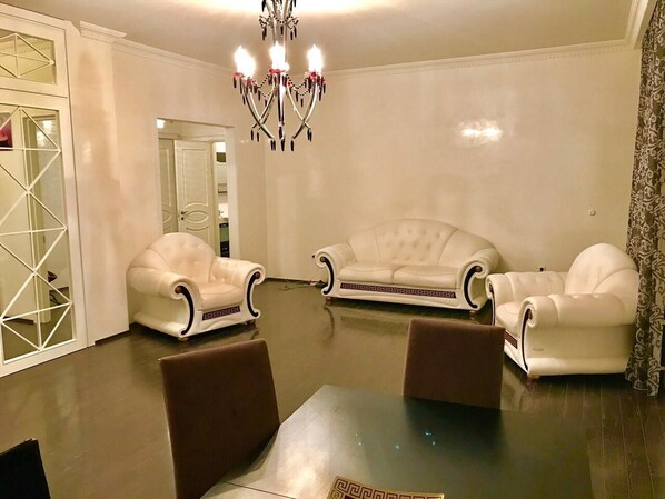 A spacious living room with a chandelier, white leather sofas, and a dining table with chairs.
