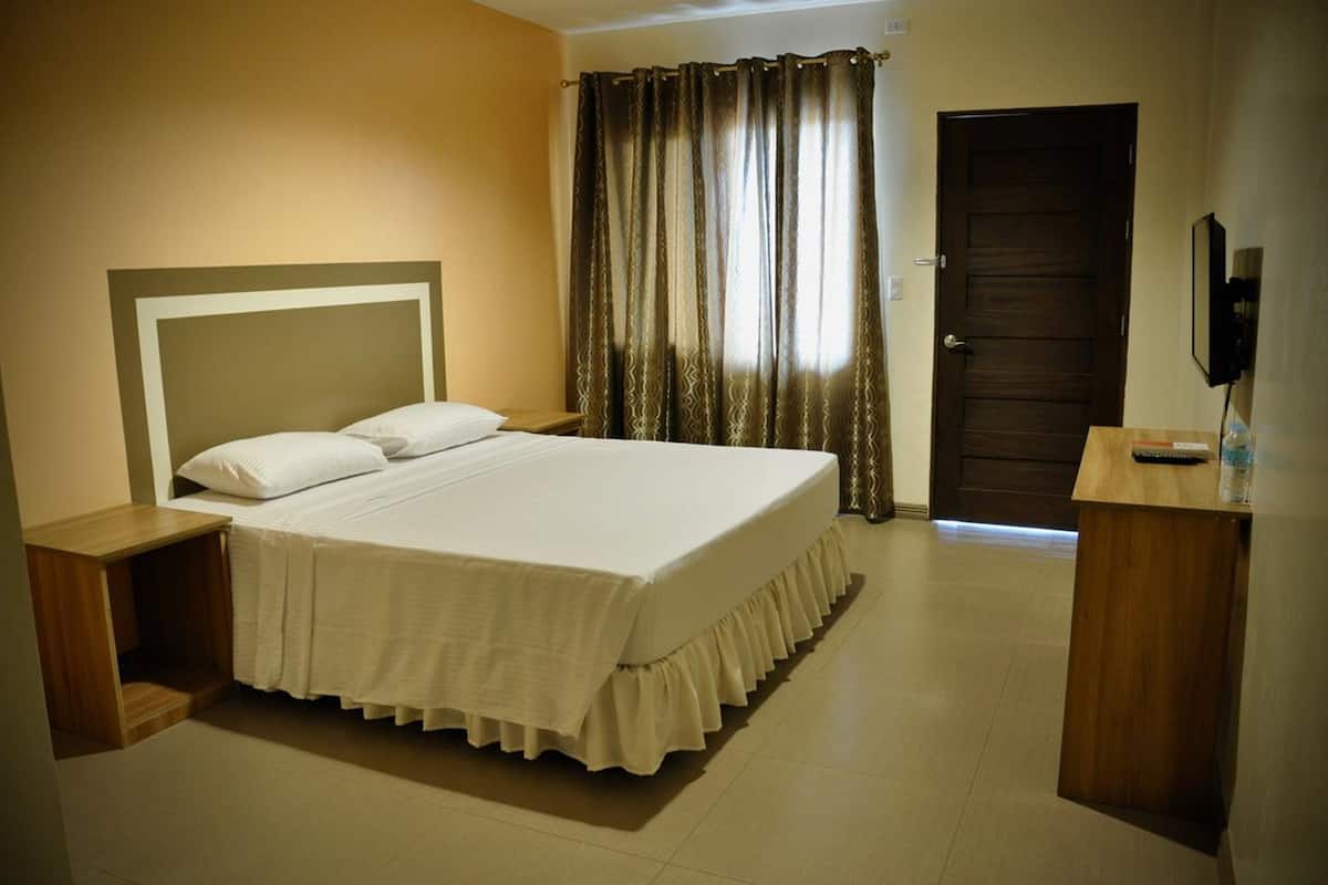 Ein ordentlich eingerichtetes Hotelzimmer mit einem großen Bett, einem Nachttisch aus Holz, einem Schreibtisch mit Stuhl und einer Tür.