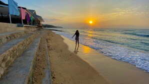Una playa con escalones que bajan al agua, una persona caminando y una puesta de sol al fondo.