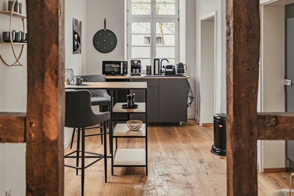 Une cuisine moderne avec un coin repas, dotée d’un plancher en bois, d’une machine à café noire, d’un four à micro-ondes et d’un porte-casseroles suspendu.