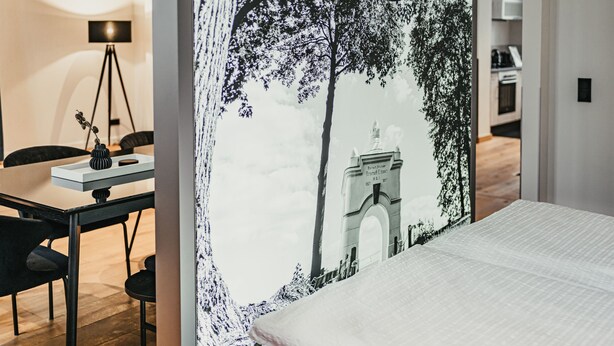 Ein Hotelzimmer mit einem Bett, einem Schreibtisch und einem Stuhl. An der Wand ist ein Wandbild von einem Baum und einem Gebäude.