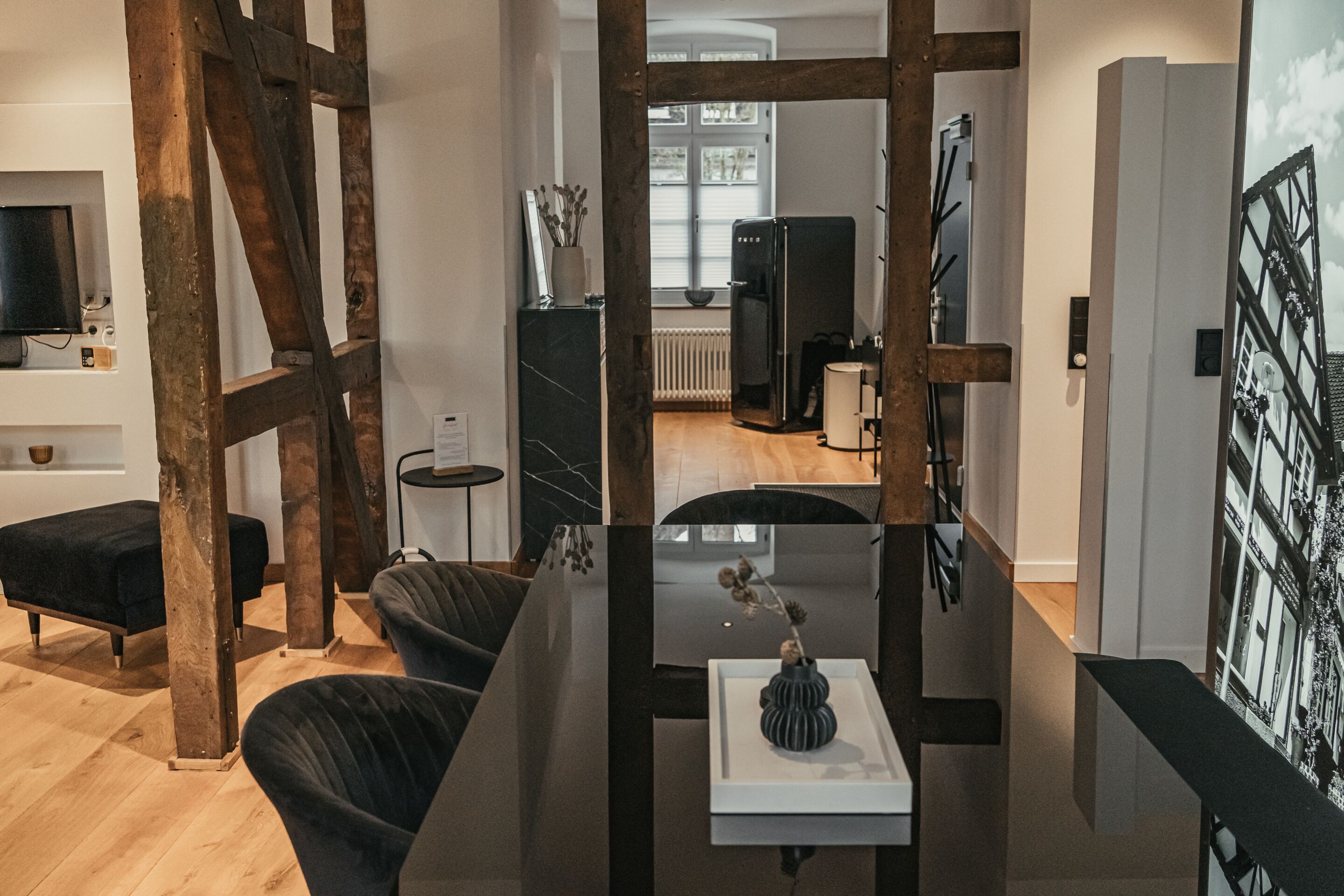 A modern living room with a wooden beam structure, a black sofa, a coffee table with a vase, and a view of a kitchen and dining area in the background.