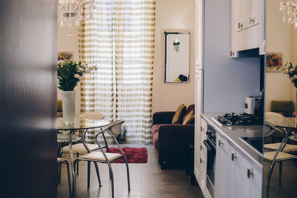 A modern kitchen with a gas stove, a dining area with a glass table and chairs, a sofa, and a red rug.