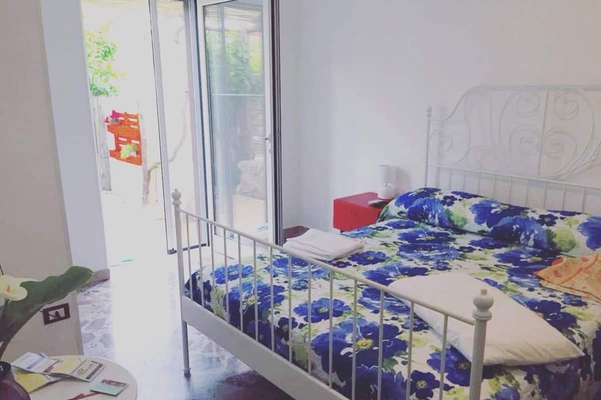 Een slaapkamer met een bed, een nachtkastje, een rode doos en een raam met uitzicht op de omgeving.