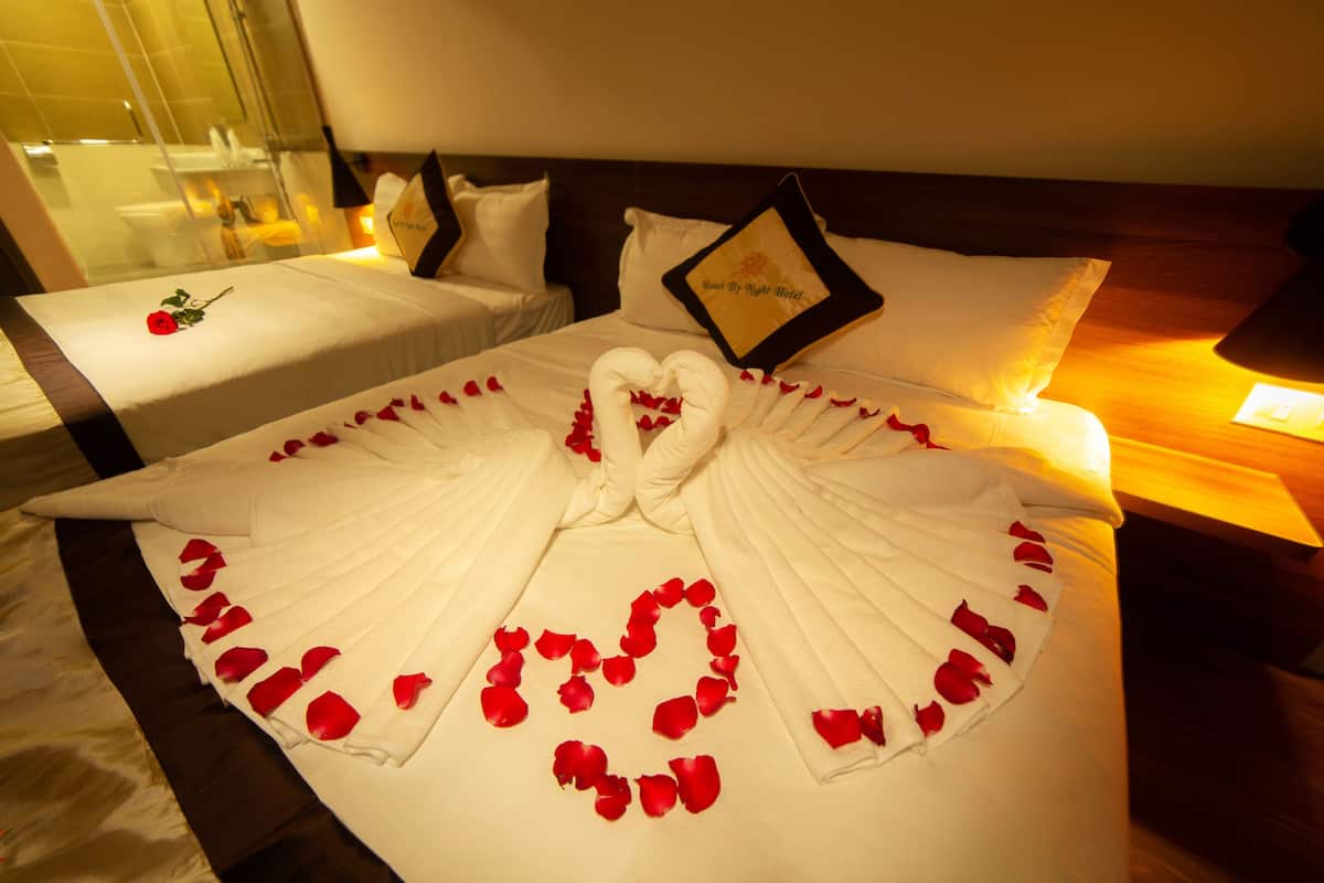 Une chambre d’hôtel avec un lit parfaitement fait, agrémenté d’une décoration en forme de cœur composée de pétales de rose et d’une sculpture réalisée avec une serviette.