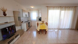Un salon avec un téléviseur, une table à manger avec des chaises, une cuisine à l’arrière-plan et une grande fenêtre avec des rideaux.