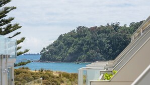 A coastal view from a modern building with a balcony overlooking a beach, a forested hill, and a distant ship. - Seaside Marine - Mt Maunganui Holiday Home (Mt Maunganui)