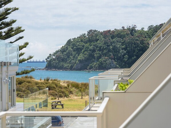 A coastal view from a modern building with a balcony overlooking a beach, a forested hill, and a distant ship.