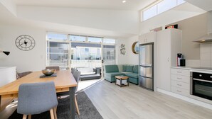 A modern kitchen with a dining area, a sofa, and a large window. - Seaside Marine - Mt Maunganui Holiday Home (Mt Maunganui)