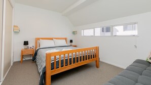 A bedroom with a wooden bed, a nightstand, a bedside table, a window with blinds, and a grey sofa. - Seaside Marine - Mt Maunganui Holiday Home (Mt Maunganui)