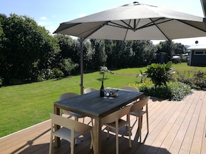 Una terraza de madera con una mesa y sillas bajo un gran paraguas, rodeada de césped y diversas plantas.